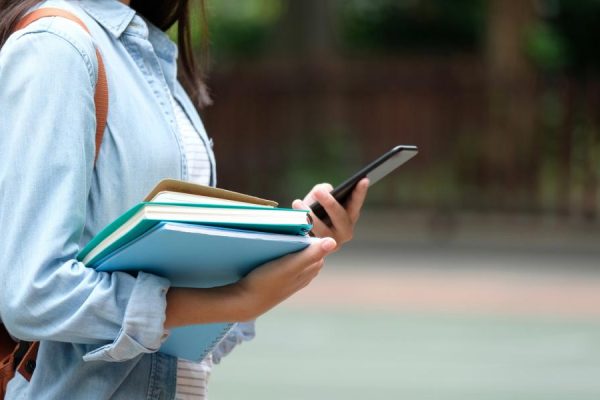 Student girl holding books and smartphone while walking in school campus background, education, back to school concept